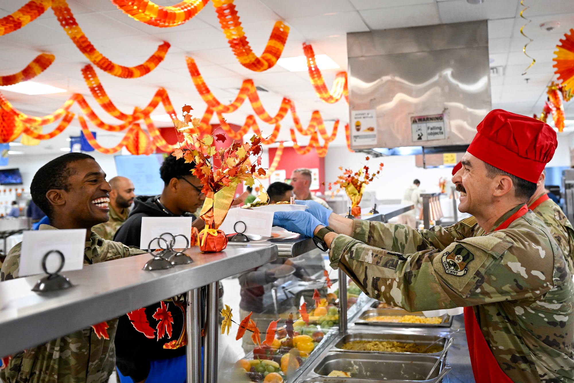 A commander wearing a red hat serves a Thanksgiving meal to an Airman in the dining facility.