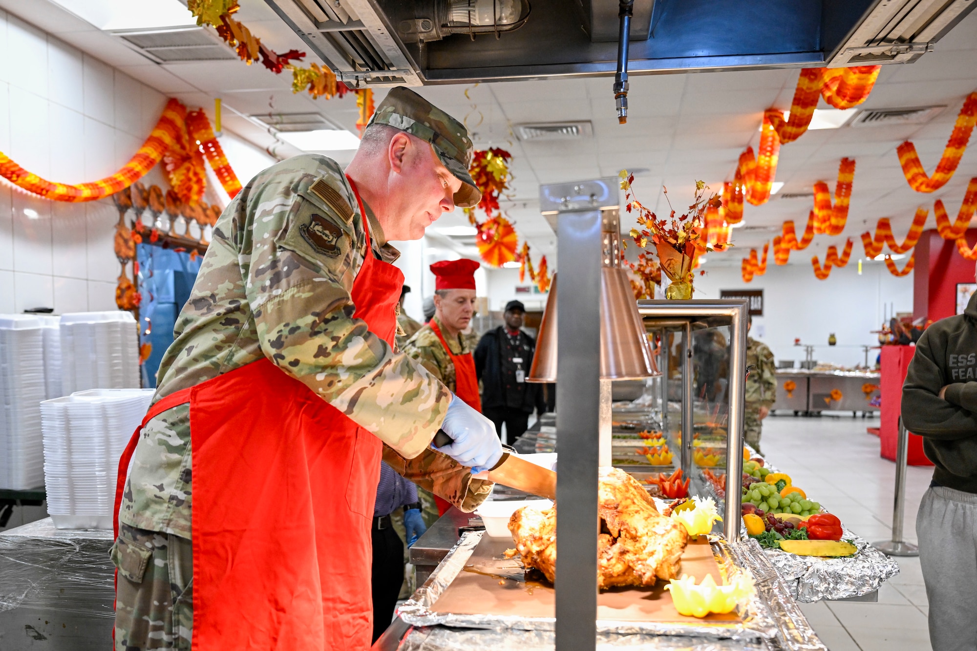 A chief master sergeant wearing a red apron carves a turkey during a Thanksgiving meal in the dining facility.