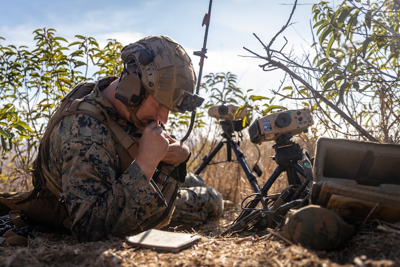 A Marine looks at a notepad on the ground while crouching next to equipment.