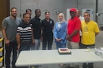 A group of nine people stand around a table with a cake on it.