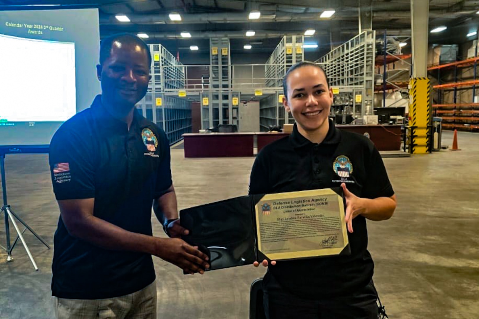 A man and woman stand in front of a projection screen in a warehouse holding a certificate.