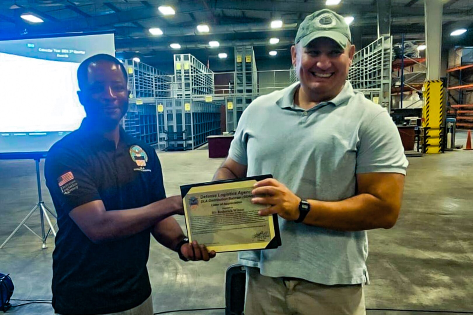 Two men stand in front of a projection screen in a warehouse shaking hands and holding a certificate.