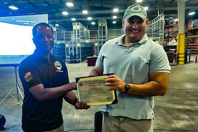 Two men stand in front of a projection screen in a warehouse shaking hands and holding a certificate.