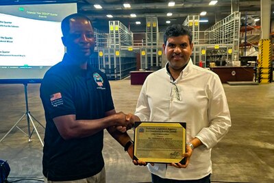 Two men stand in front of a projection screen in a warehouse shaking hands and holding a certificate.