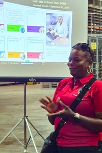 A woman in a red shirt stands in front of a projection screen speaking.
