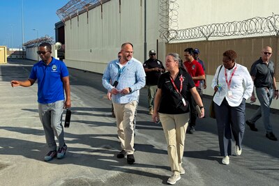 A group of people walk outside on a sunny day in front of buildings and protective fencing.