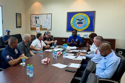 A group of people sit around a conference table talking. There is a framed logo on the back wall that says "DLA Distribution Bahrain Southwest Asia" with the DLA logo on it.