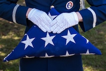 A Honorguardsman wearing a blue dress uniform with white accents and badges, holds a folded U.S. Flag with the stars showing.