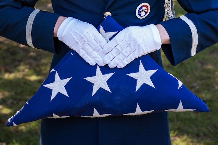 A Honorguardsman wearing a blue dress uniform with white accents and badges, holds a folded U.S. Flag with the stars showing.