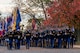 Several rows of Soldiers dressed in dark blue ceremonial dress uniforms are marching down a street (toward the camera). The Soldiers on the left are carrying flags and the Soldiers on the right are carrying rifles.