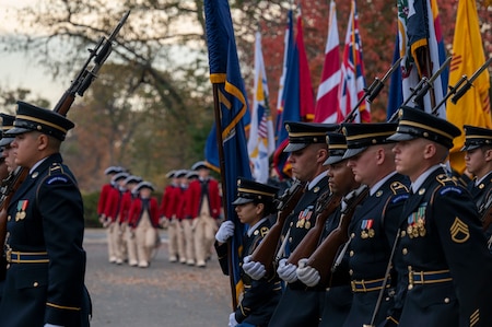 Soldiers dressed in dark blue ceremonial dress uniforms are marching down a street (to the left), and in the distance is another group of Soldiers dressed in red Revolutionary War-era uniforms