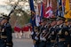 Soldiers dressed in dark blue ceremonial dress uniforms are marching down a street (to the left), and in the distance is another group of Soldiers dressed in red Revolutionary War-era uniforms