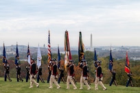 Soldiers in blue Revolutionary War era uniforms march across a green lawn while carrying various flags.