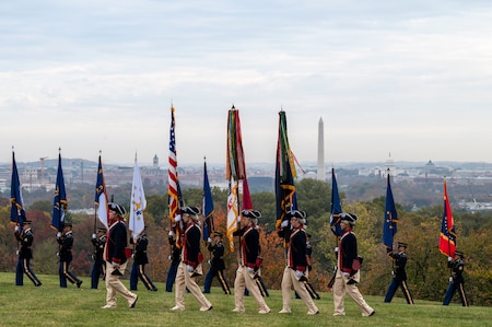 Soldiers in blue Revolutionary War era uniforms march across a green lawn while carrying various flags.