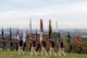 Soldiers in blue Revolutionary War era uniforms march across a green lawn while carrying various flags.