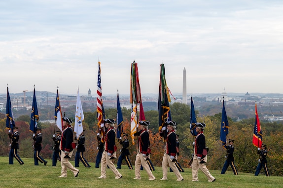 Soldiers in blue Revolutionary War era uniforms march across a green lawn while carrying various flags.
