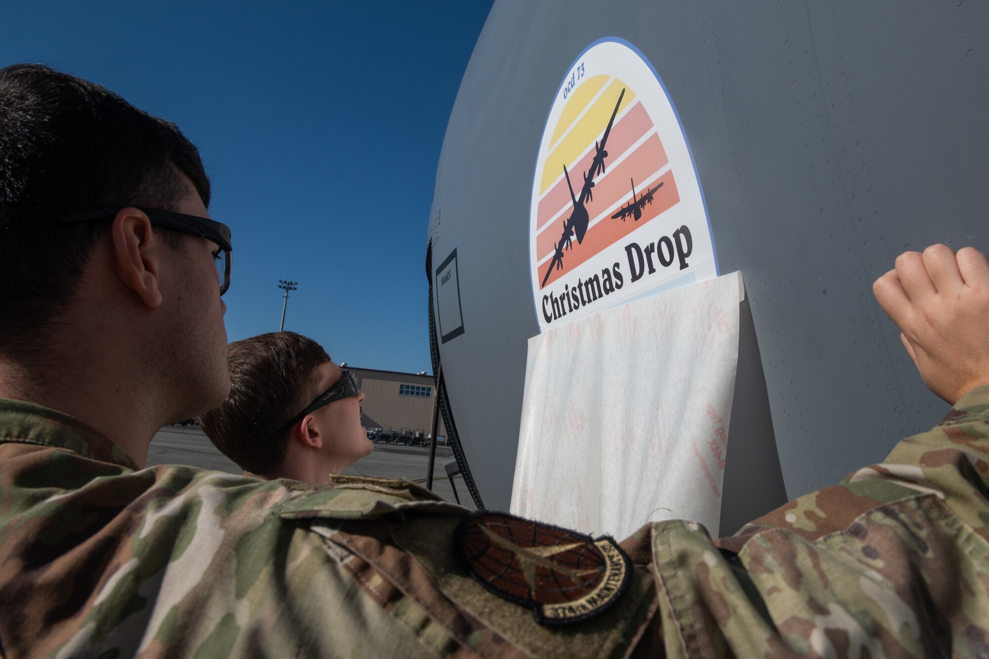 Two airmen peel a backing off of the Operation Christmas Drop while sticking it to an aircraft.