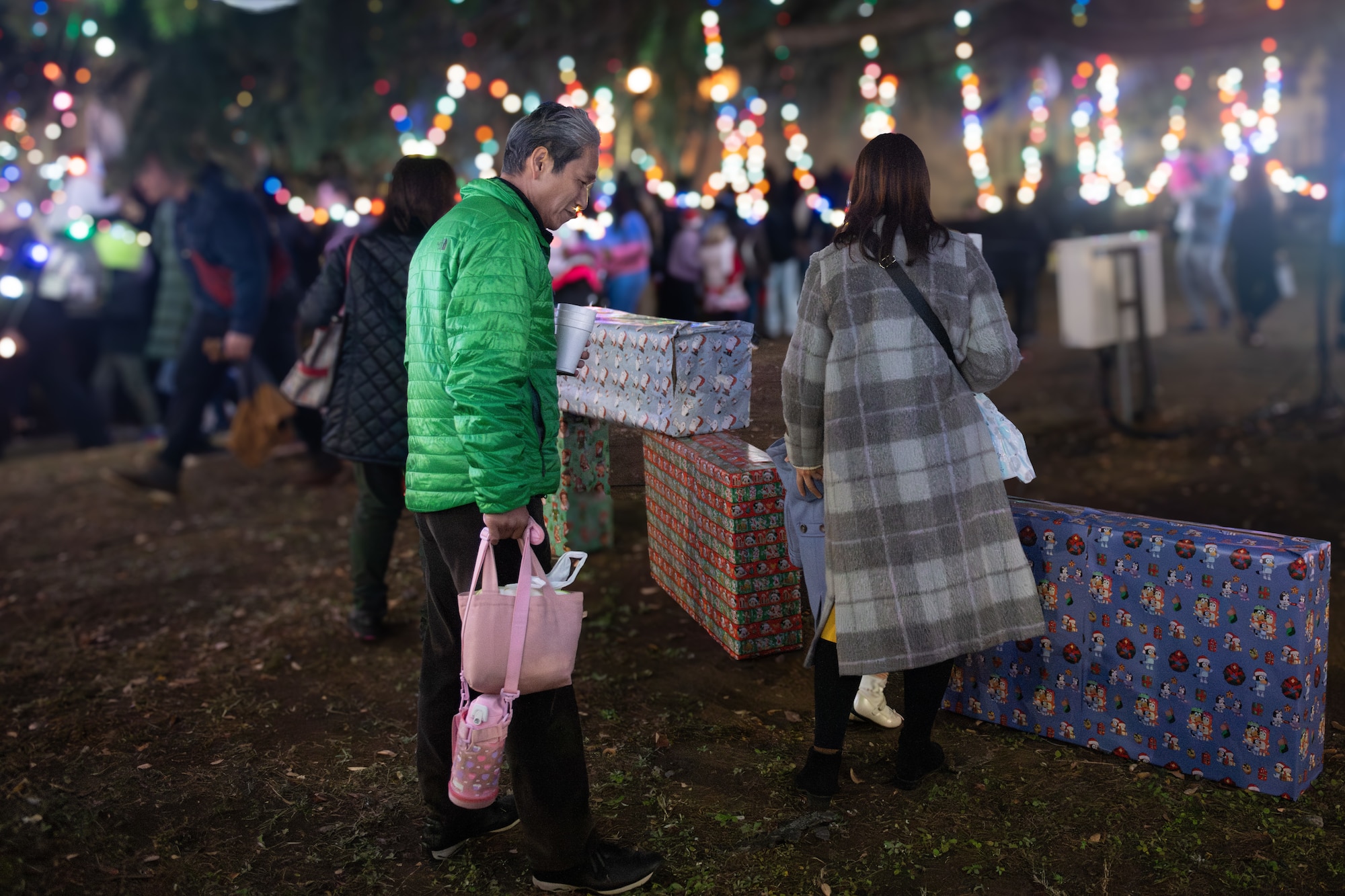 Attendees of the tree lighting ceremony walk toward the tree and interact with the prop gift boxes lying on the ground.