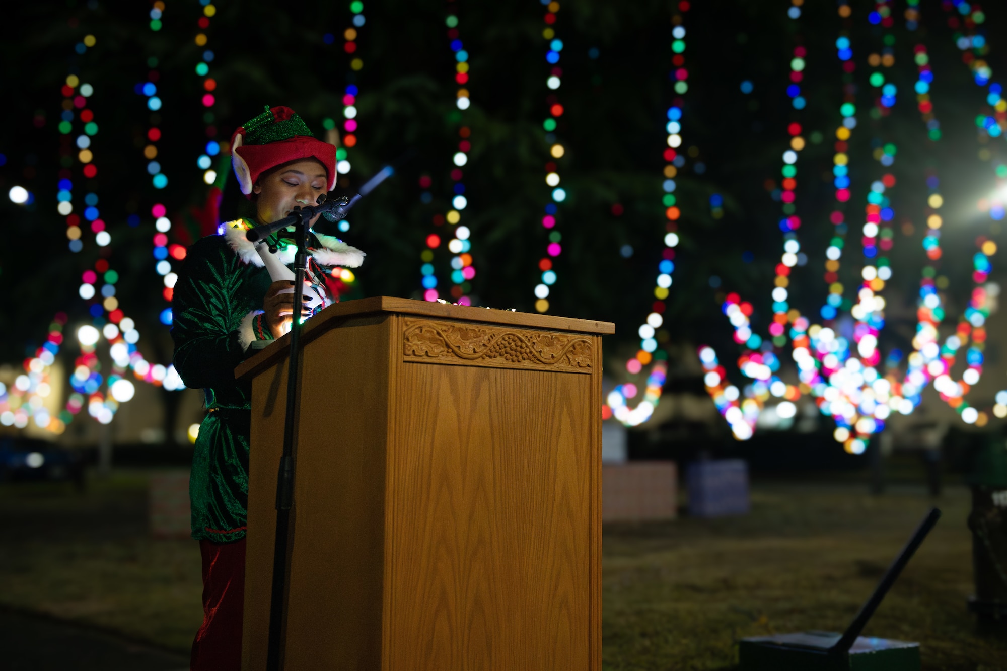 A volunteer dressed in a green and red elf costume speaks to a crowd from a wooden podium.