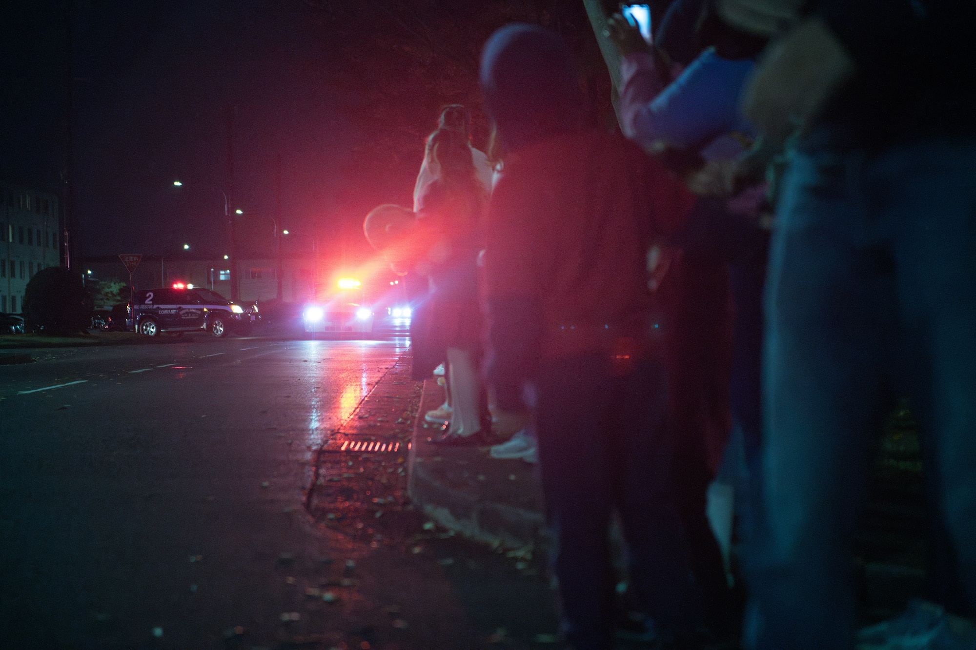 People watch alongside the sidewalk as emergency vehicles drive down the road toward them