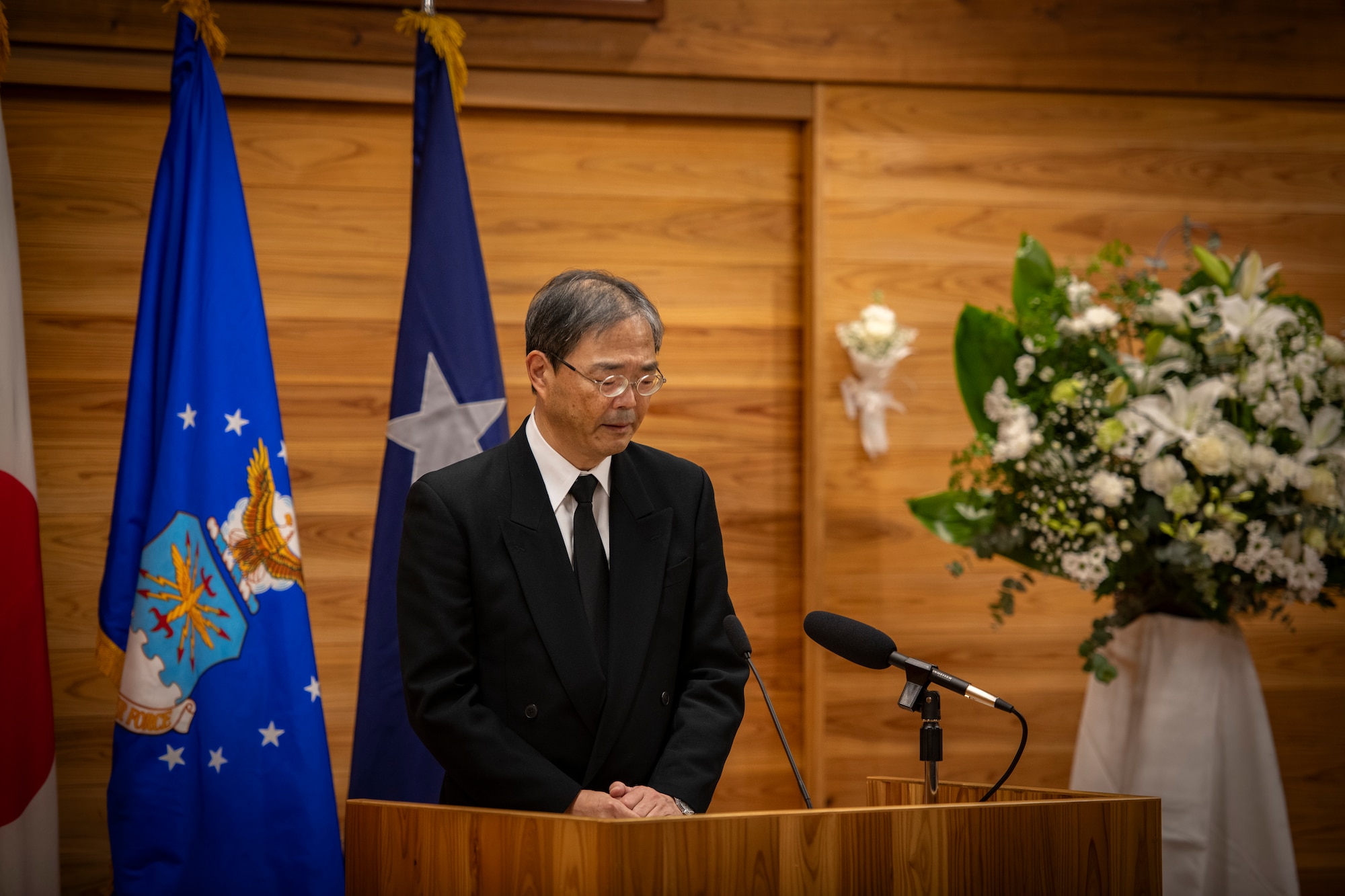 A man stands at a lectern and speaks into a microphone in front of three flags.