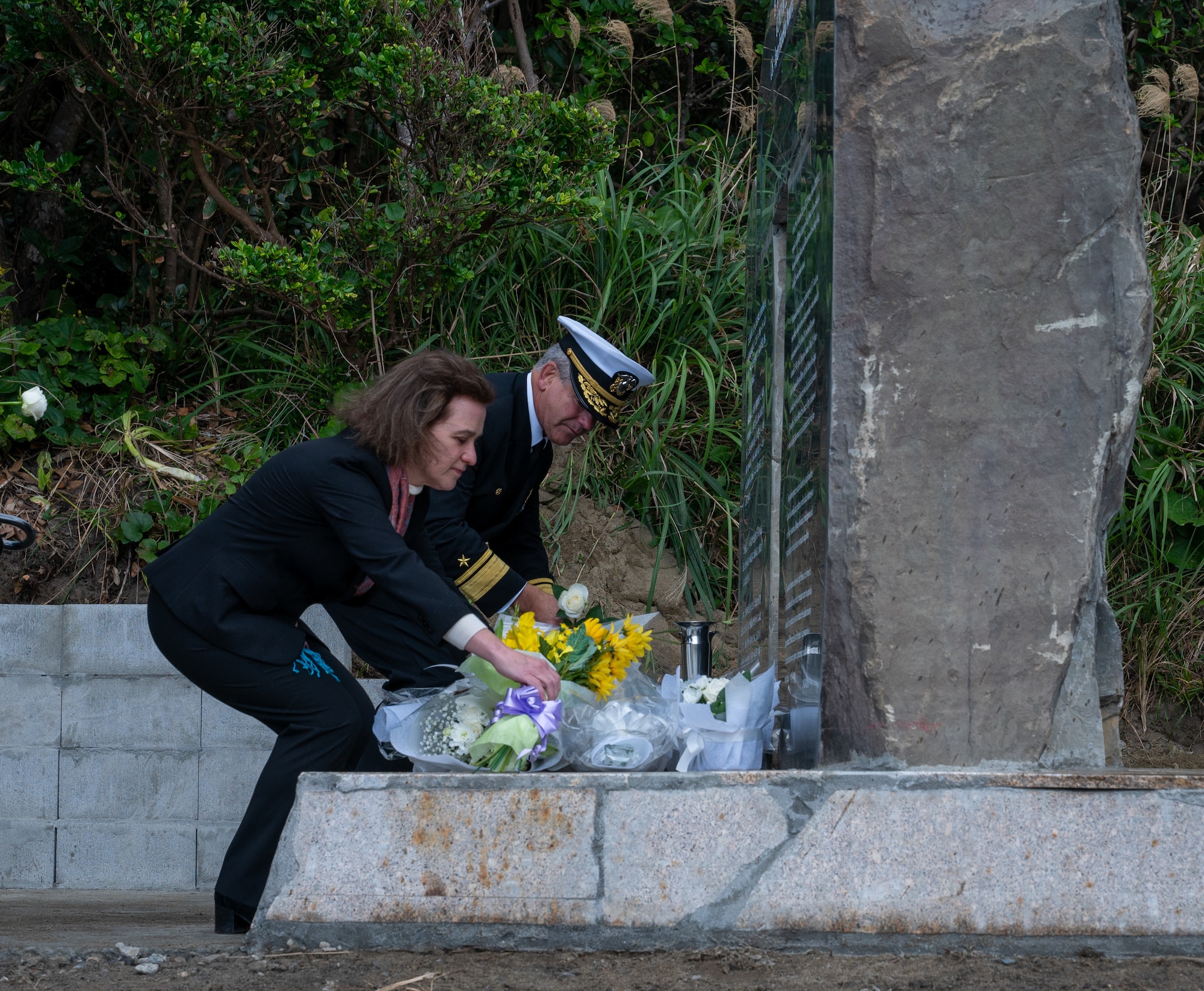 Two people are bending down placing bouquets of flowers on the base of a stone monument.