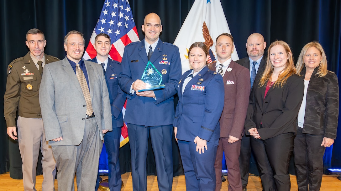 Nine individuals in military uniforms and business attire stand near each other on a stage. One man in the middle holds an award.