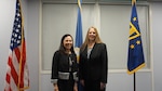 Two professionally dressed people stand in the center of three flags against a white wall.