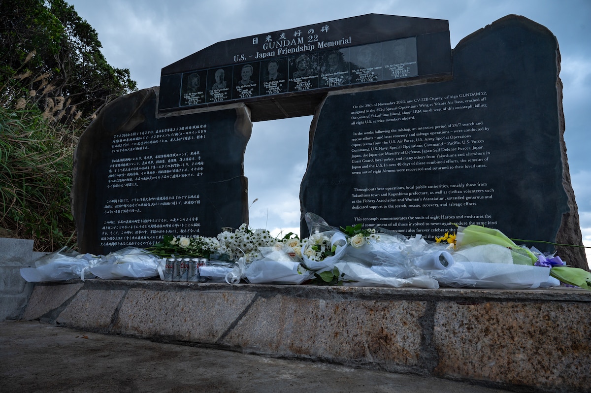 Stone monument stands on concrete/stone base with flowers and offerings resting on the base.
