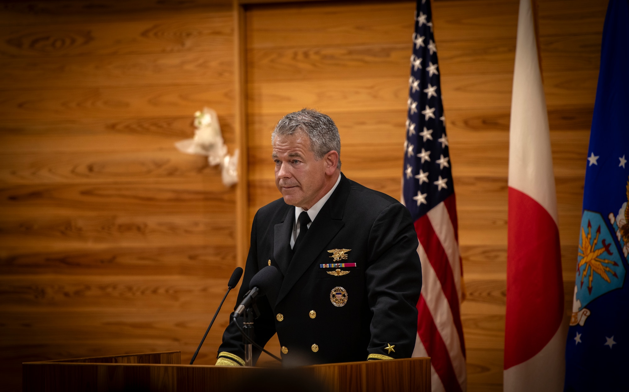 A man stands behind a lectern while speaking into a microphone in front of three flags.