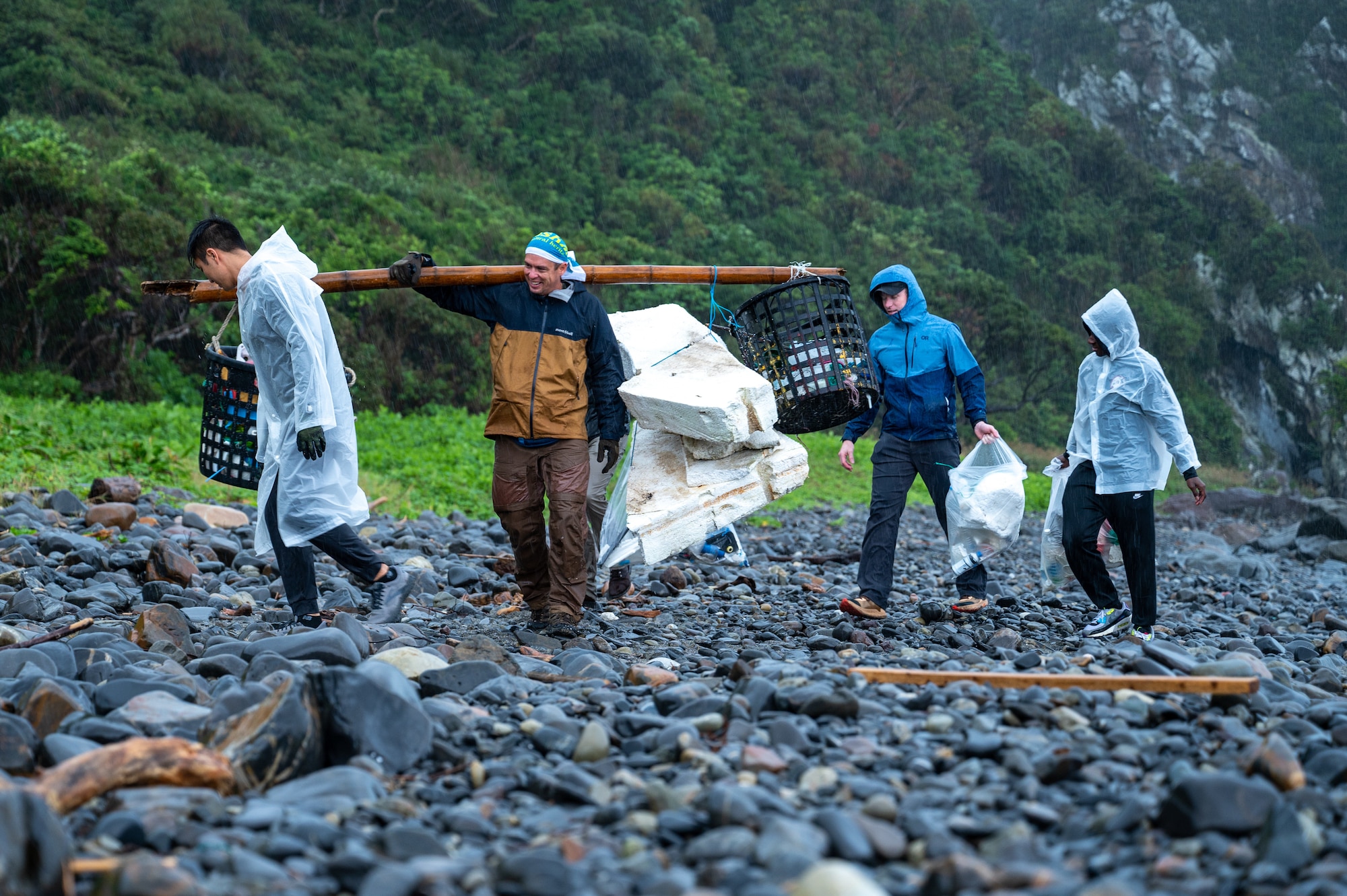Four men walk in a line across a rocky shore carrying garbage.