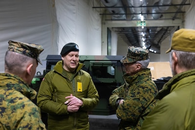 Four men in green camo uniforms stand in what appears to be a warehouse talking