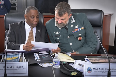 A black man in a suit and a white man in military dress uniform look over papers at a desk.