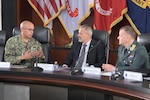 Three men, two in military uniform, sit at a desk talking.