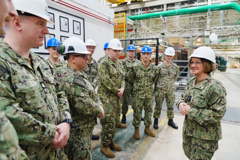 A woman leader in military uniform speaks to a group of service members in a warehouse.