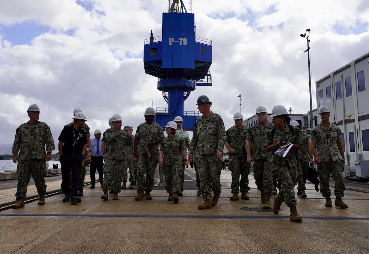A group of service members in uniform walk through a shipyard.