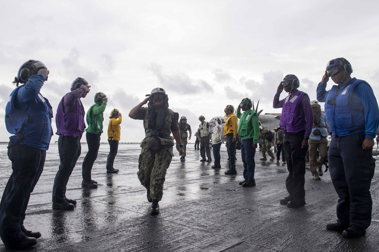 Service members in colored uniforms and flight gear render a salute to a female military leader as she walks through the flight deck aboard a Navy ship.