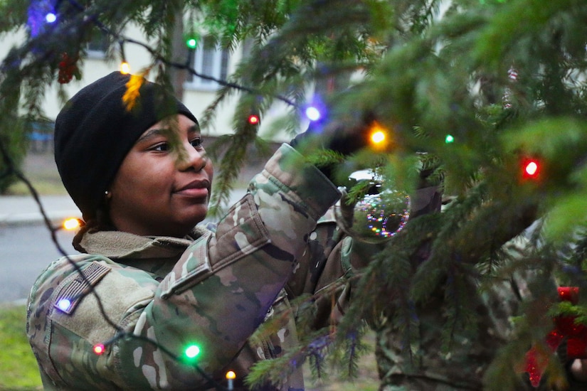 A soldier hangs an ornament on a Christmas tree with colorful lights.