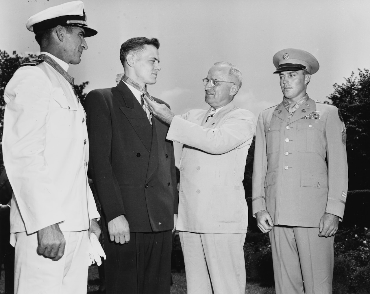 Four men stand together. One places a medal over another of the men’s necks.