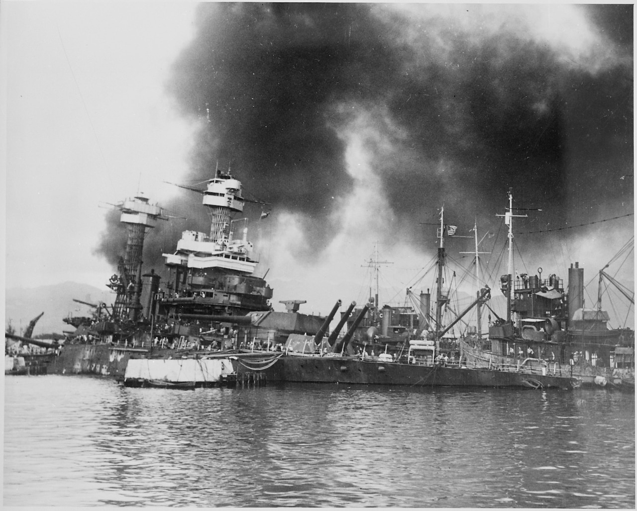 A large ship lists slightly to the side as heavy smoke plumes off it.