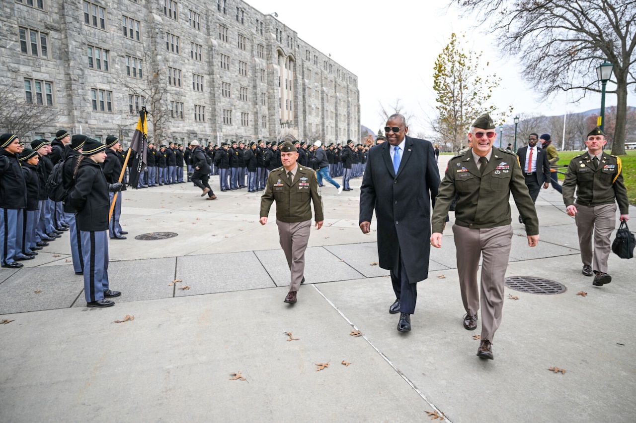 A man in business attire walks between service members in uniform.