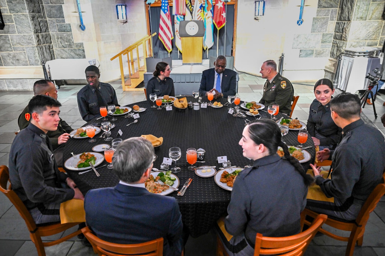 A group of military academy cadets and a man in business attire are seated around a table.