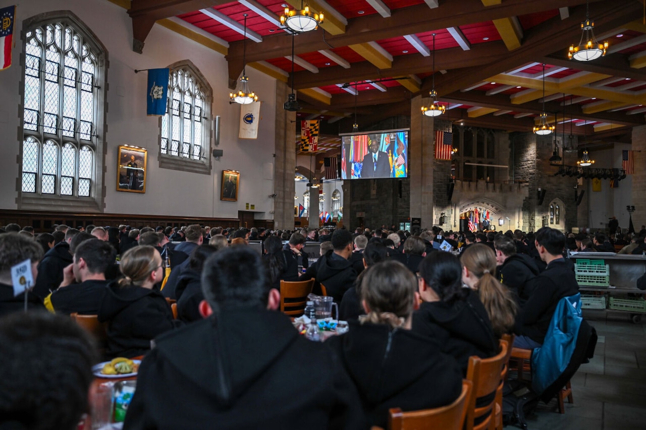A group of military academy cadets are seated at rows of tables.