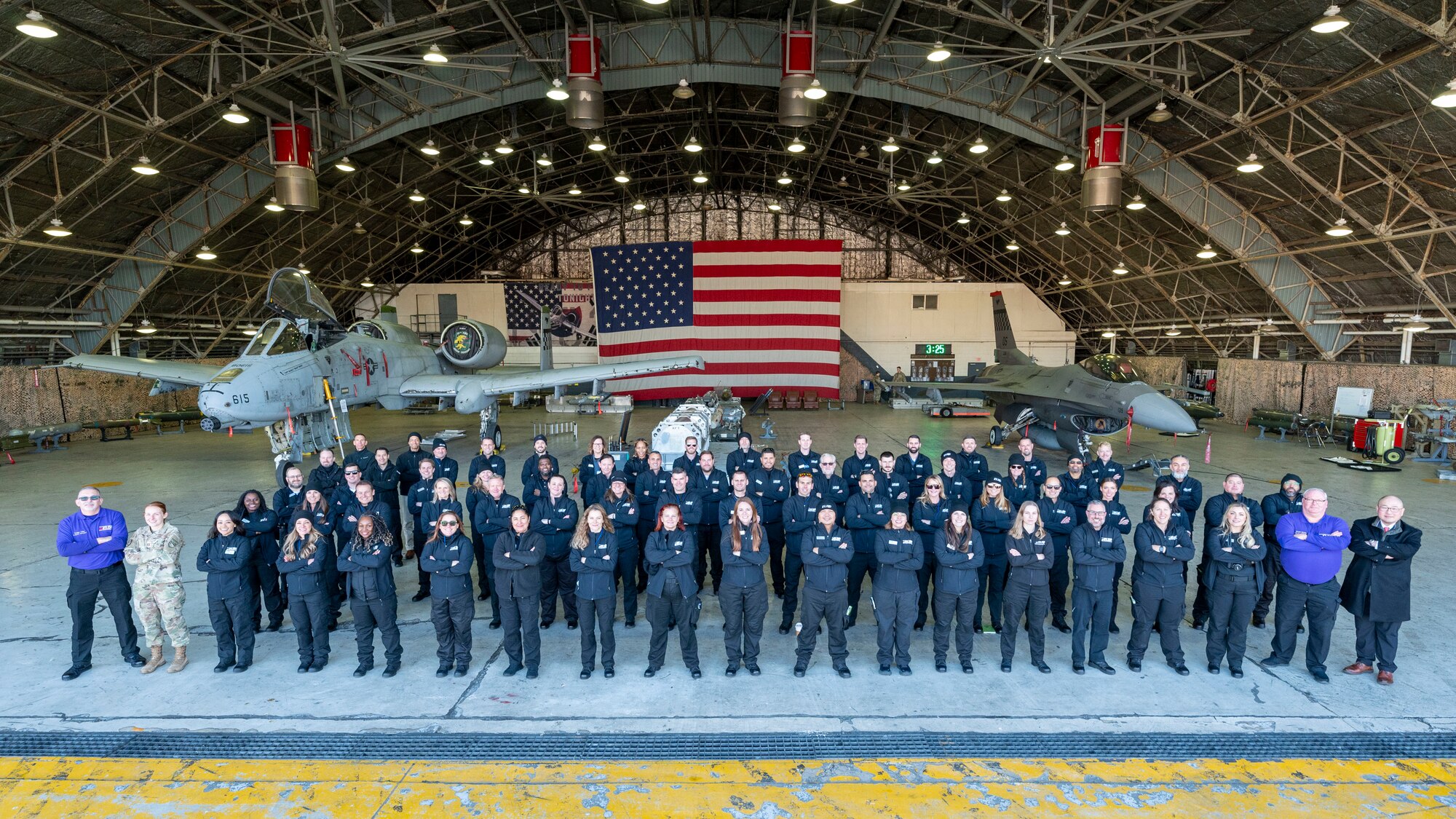 A group of people stand in formation in front of an A-10 Warthog and an F-16 Fighting Falcon in an aircraft hangar.