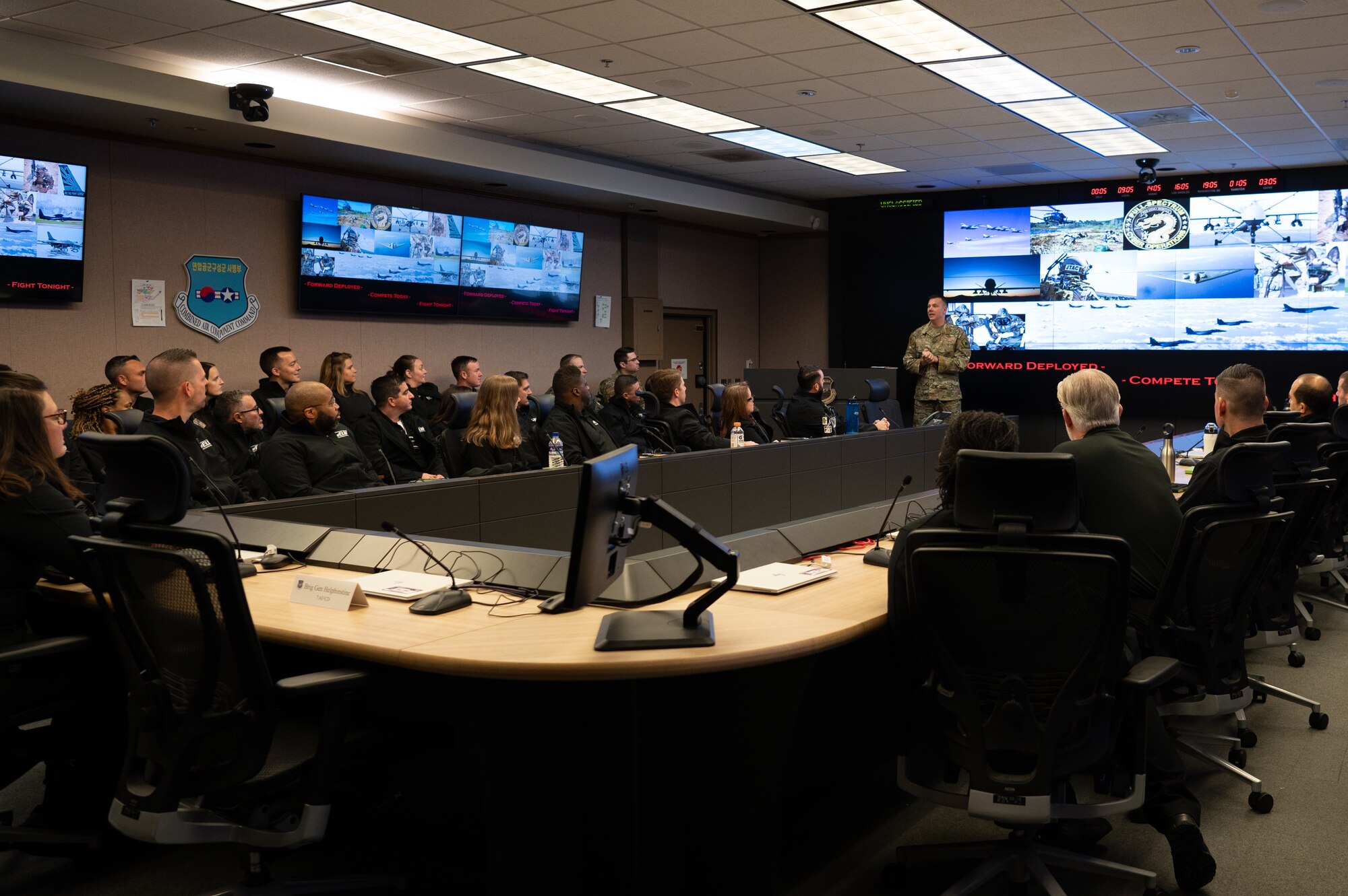 A group of people in all black sit in a conference as a man in OCPs talks at the front of the room. There is a photo collage on the screens around the room.