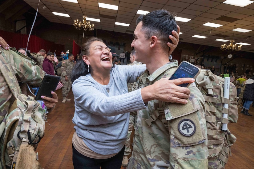 A civilian laughs while greeting a soldier wearing a backpack. Lots of other soldiers and civilians stand in the background in a large room.