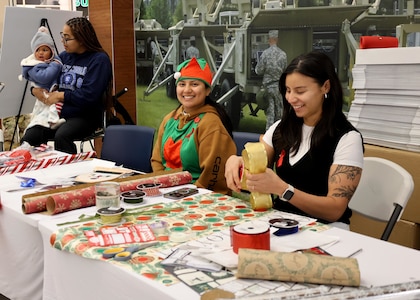 Two women sit at a table with holiday gift wrap waiting to wrap presents while another woman sits in the background holding a baby.