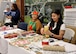 Two women sit at a table with holiday gift wrap waiting to wrap presents while another woman sits in the background holding a baby.