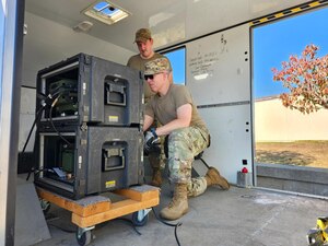 Two Airmen work on communications equipment inside of a trailer.