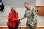 A black woman in a red jacket and white man in camo uniform shake hands in front of a stage.